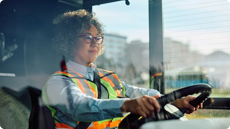 A woman wearing eye glasses, seated in the driver's seat of a bus, holding the steering wheel with both hands and looking straight ahead.