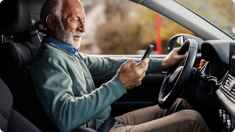 A man with a white beard and hair sitting in the driver's seat of a car, he is looking down at a smartphone he is holding in his hands.
