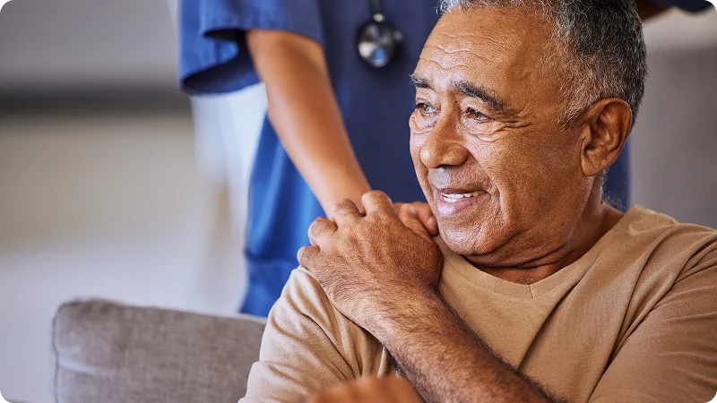 A smiling, elderly man sitting down while a nurse stands behind him with a hand on his shoulder.