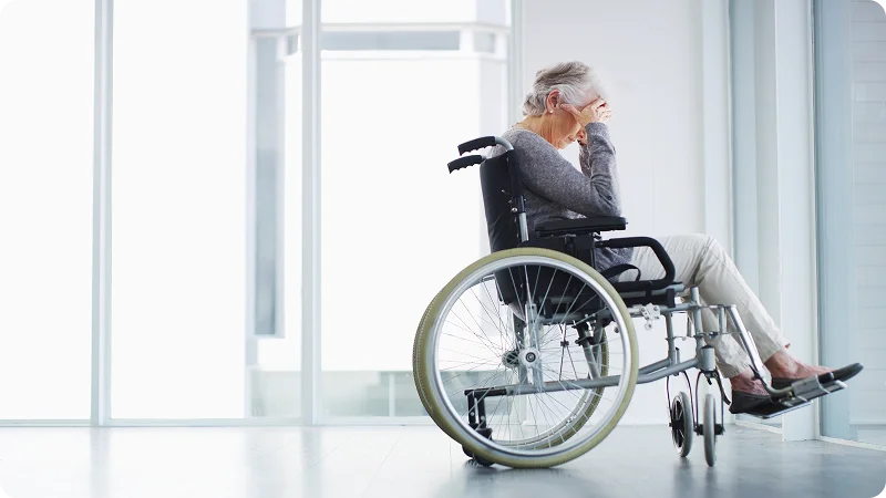 An elderly woman sitting in a wheelchair, with her head in her hands, looking distressed.