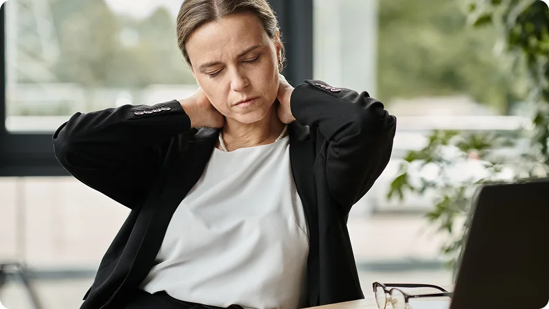 A woman, sitting at a desk and holding the back of her neck with both hands, a discomfort or pain possibly from an injury, stress, or muscle strain.