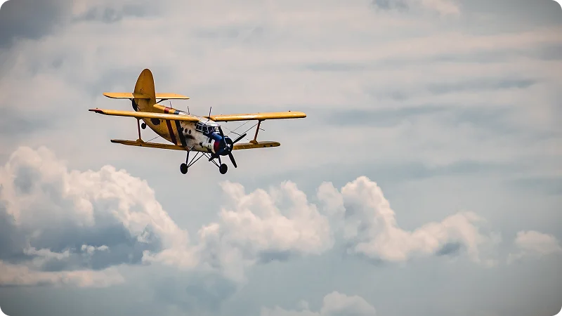 A small, yellow and white biplane flying through a cloudy sky.