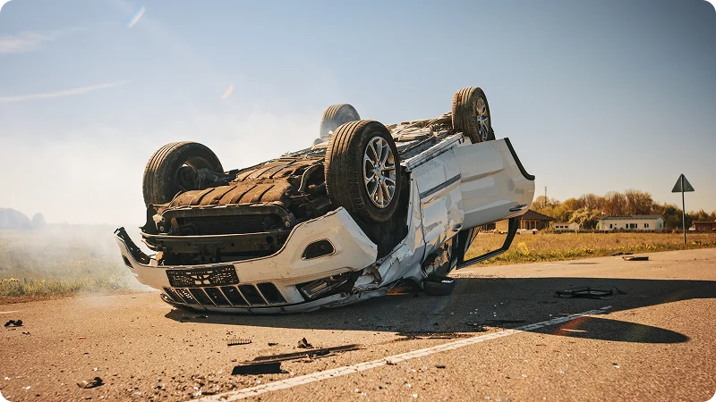 A white car crashed and it's lying upside down on a road and it is crumpled.
