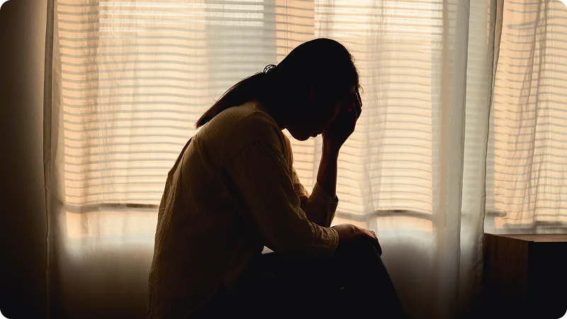 A person sitting in front of a window, with their head in their hand, looking down.