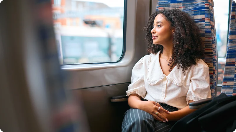 A young woman with curly hair sitting on a train, looking out the window with a thoughtful or serene expression.