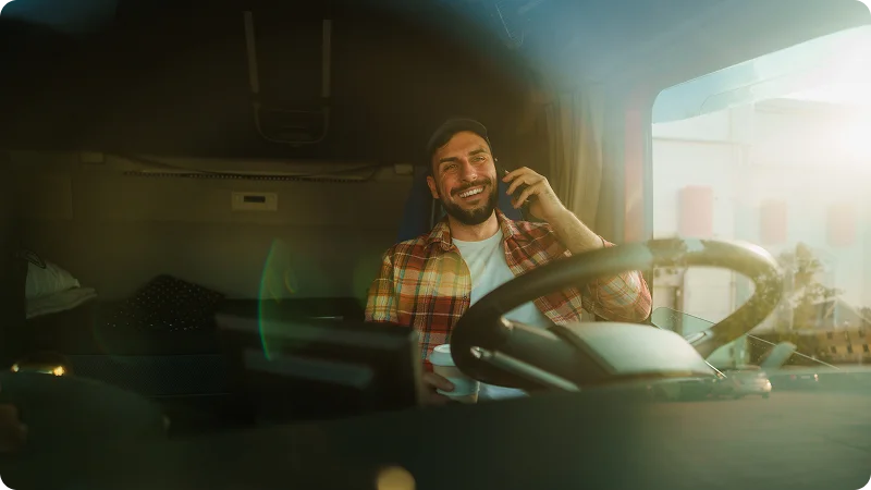 A male truck driver sitting in the cab of his truck, smiling and holding a mobile phone to his ear.