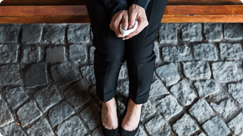 A woman sit on a bench with their hands clasped together, holding a small, white object.